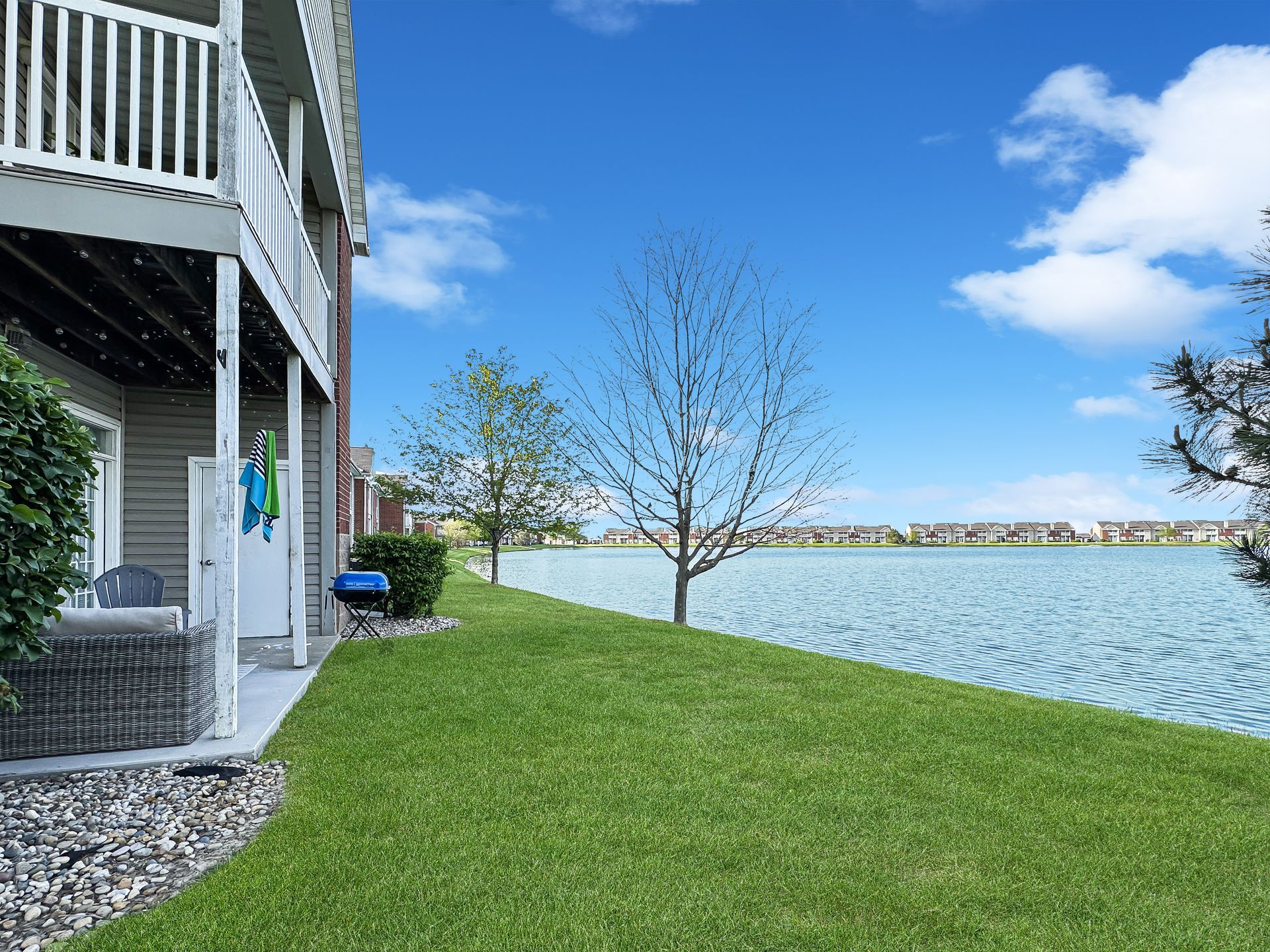 A lakeside home with a green lawn, clear blue sky, and a grill near the patio.