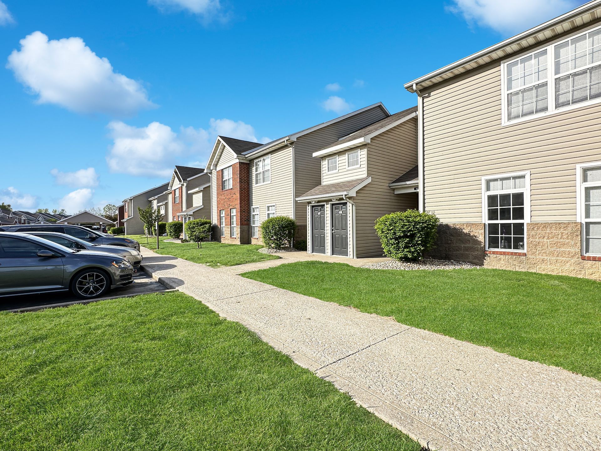 Apartment complex with row of buildings, stone walkway, cars parked along the side, blue sky.
