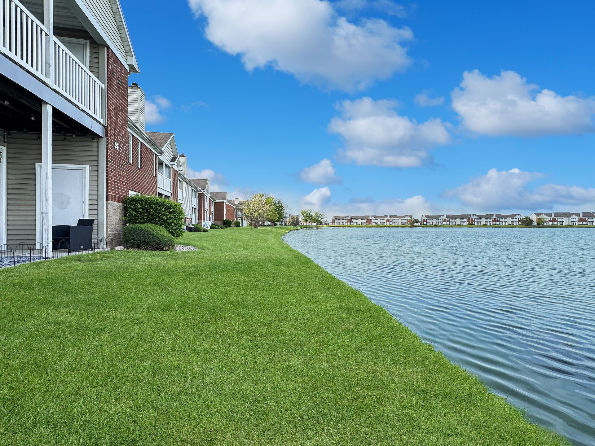 Apartment buildings with balconies overlook a grassy lawn and a lake on a sunny day.