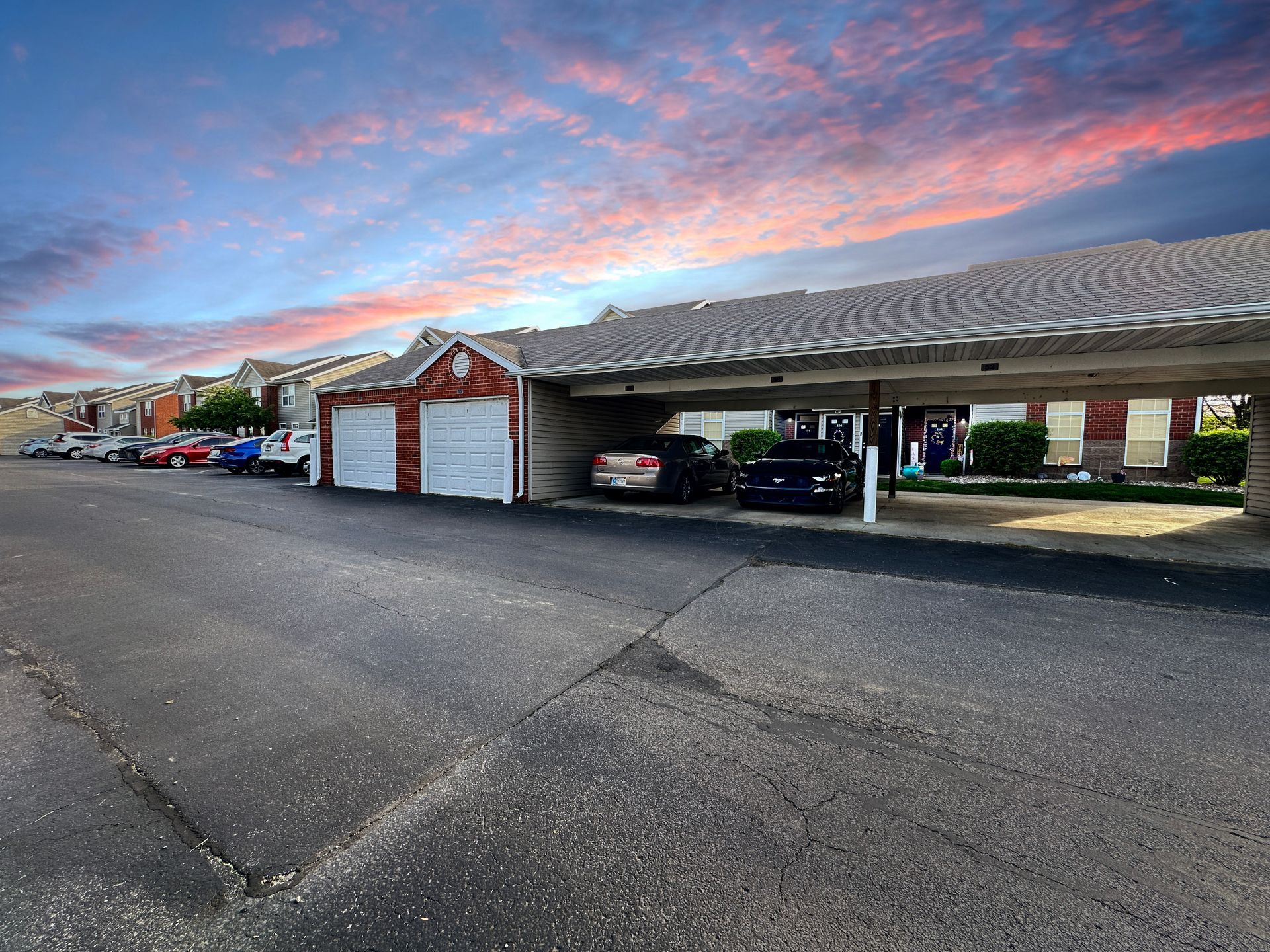 Apartment complex exterior with cars parked under a covered area, sunset sky.