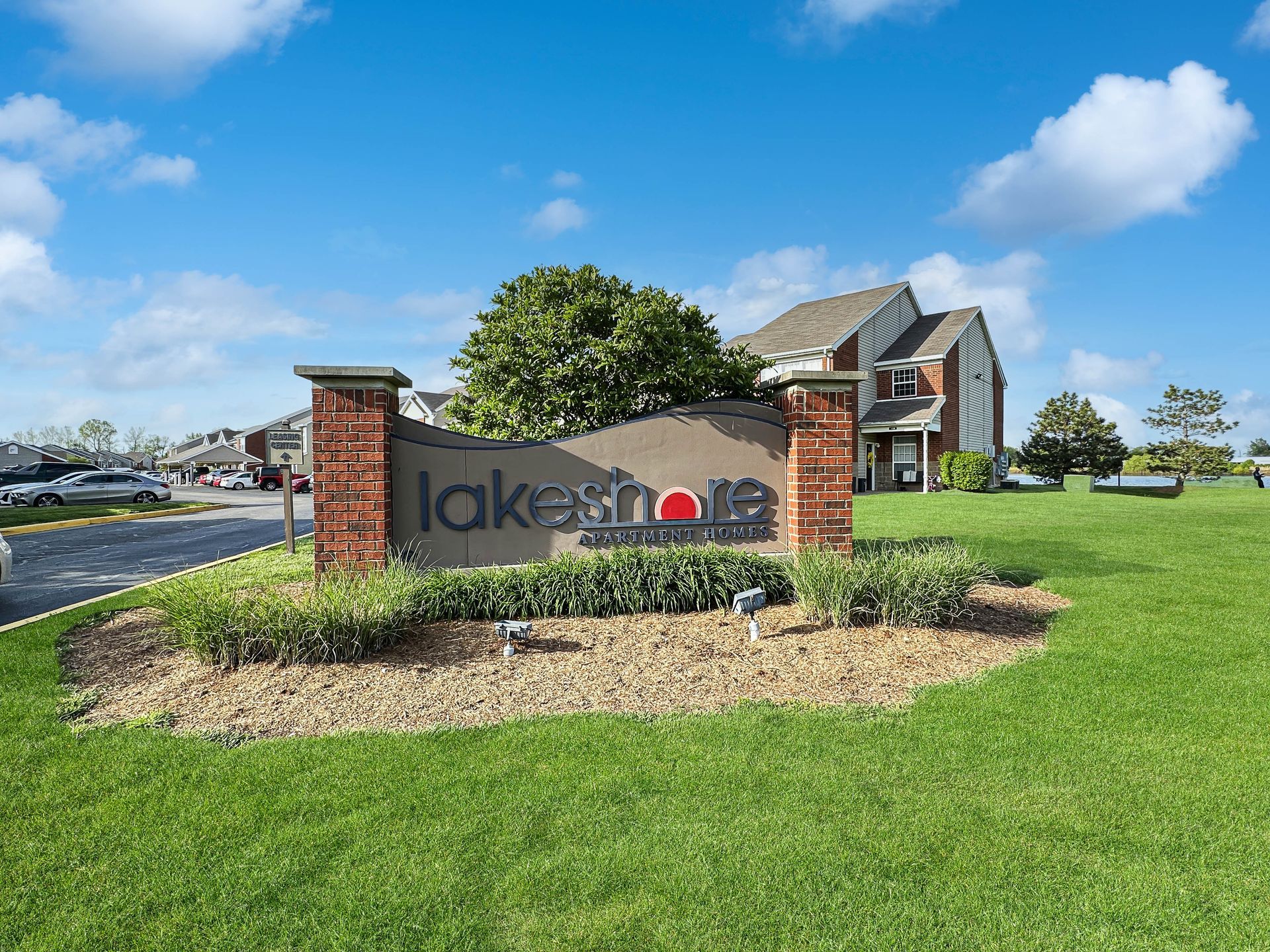Lakesmile Apartment sign with red brick accents, green grass, and blue sky background.