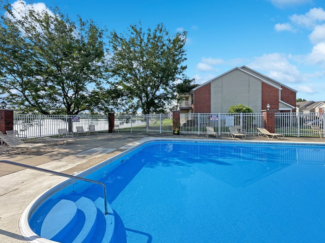 Pool with blue water and white steps, near a building and trees on a sunny day.