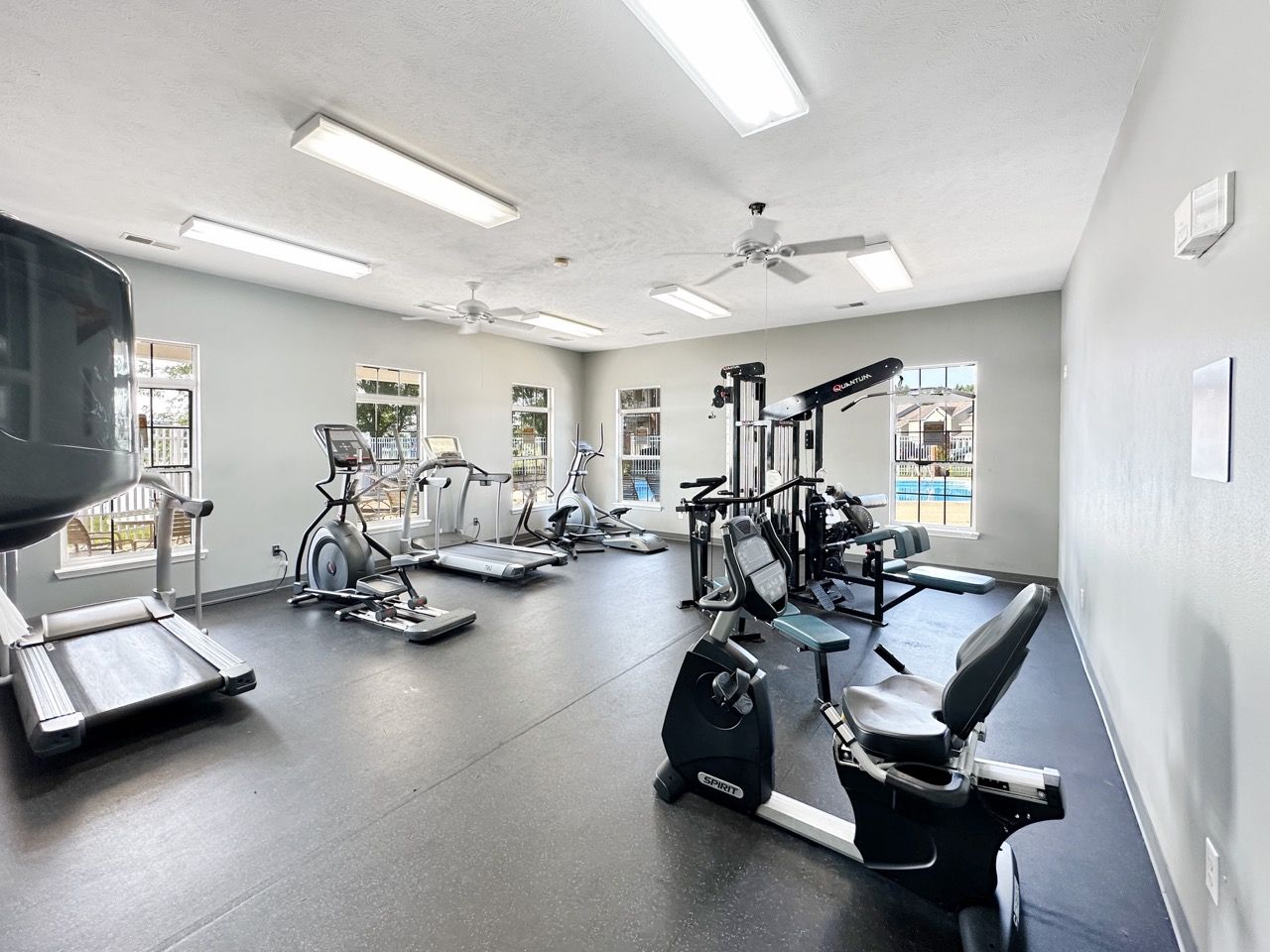 Gym with treadmills, bikes, and weights. Gray walls, black floor, and natural light from windows.