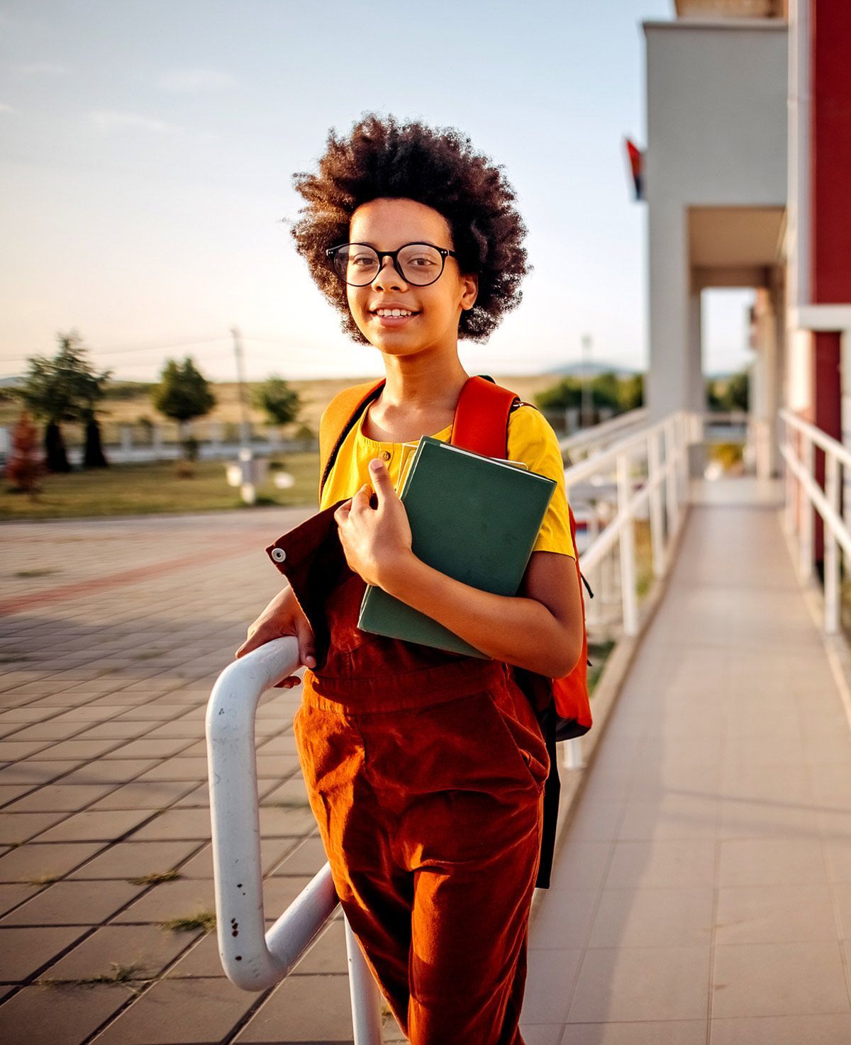 A young girl is walking down a sidewalk holding a book and a backpack