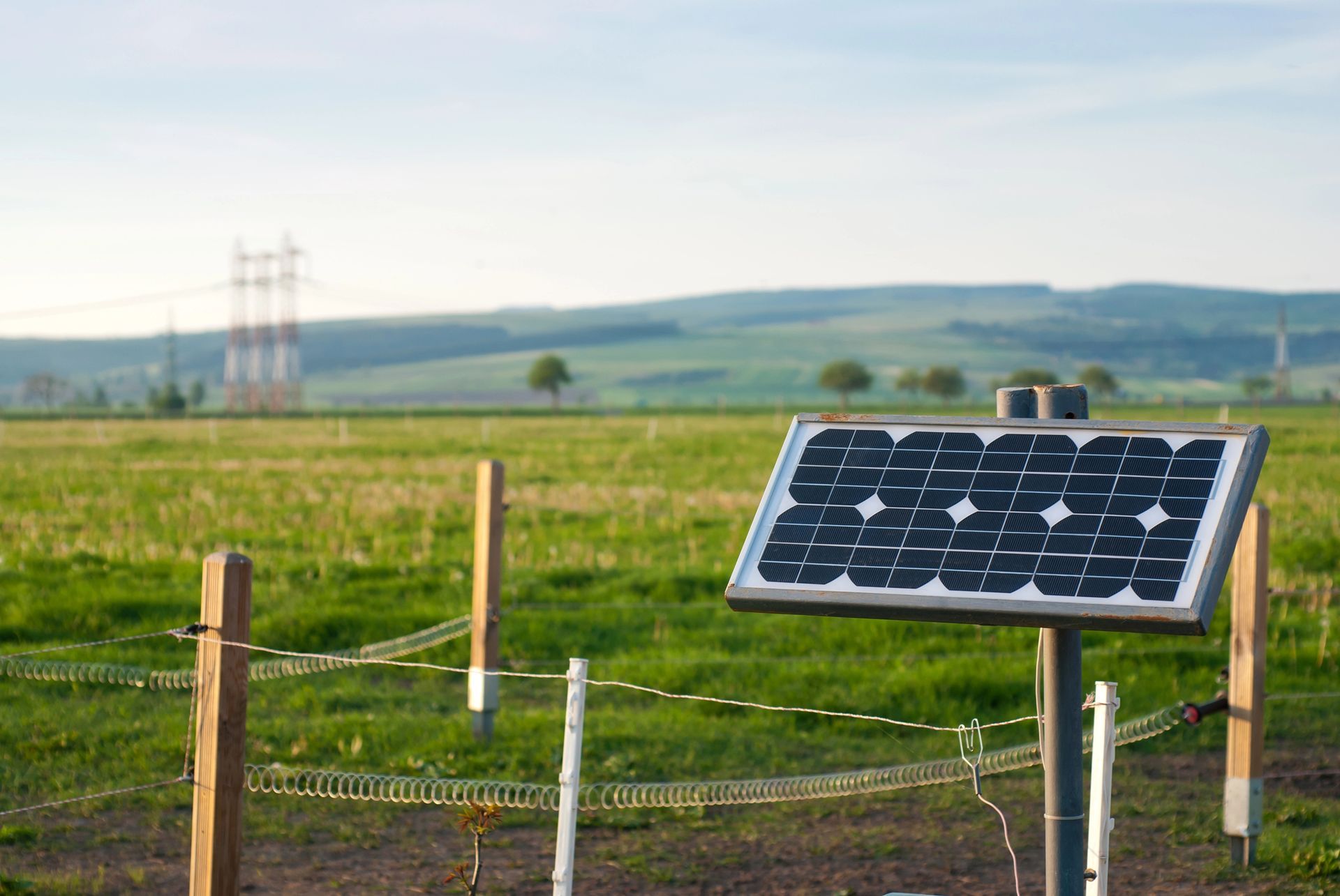 Panel solar que alimenta una cerca eléctrica en un campo verde con colinas onduladas y líneas eléctricas en la distancia.