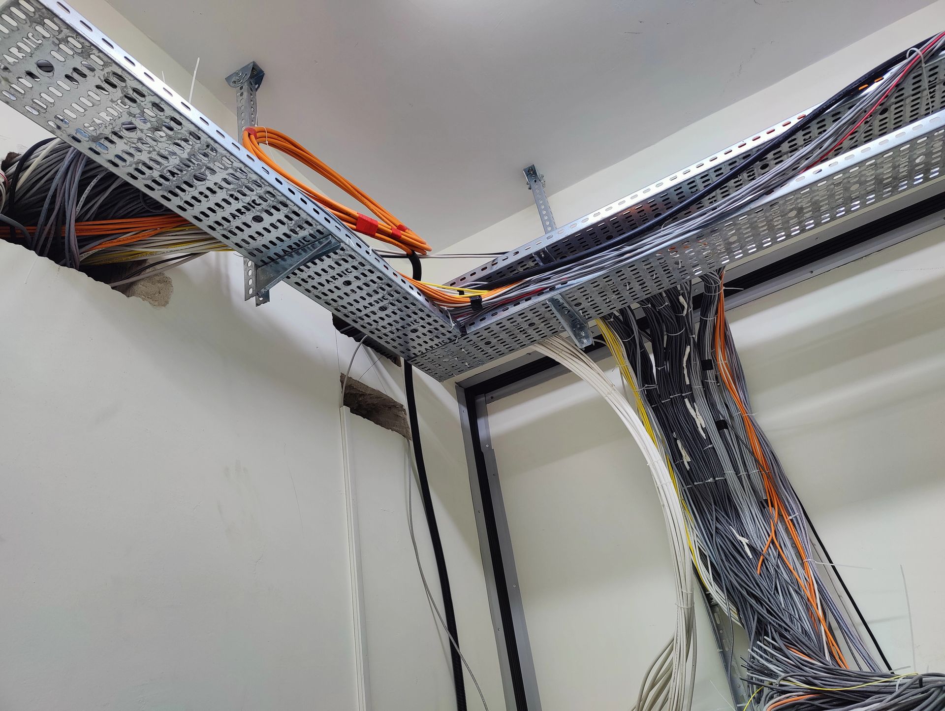 Bandejas de cables metálicas suspendidas en el aire que transportan haces de cables a través de una esquina de una habitación, montadas en una pared blanca.