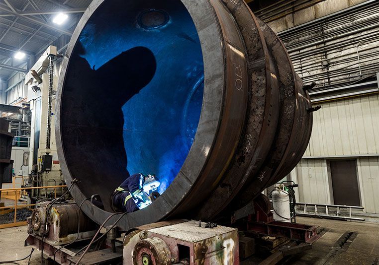 Welder wearing protective gear, working on a red metal structure in a workshop setting.