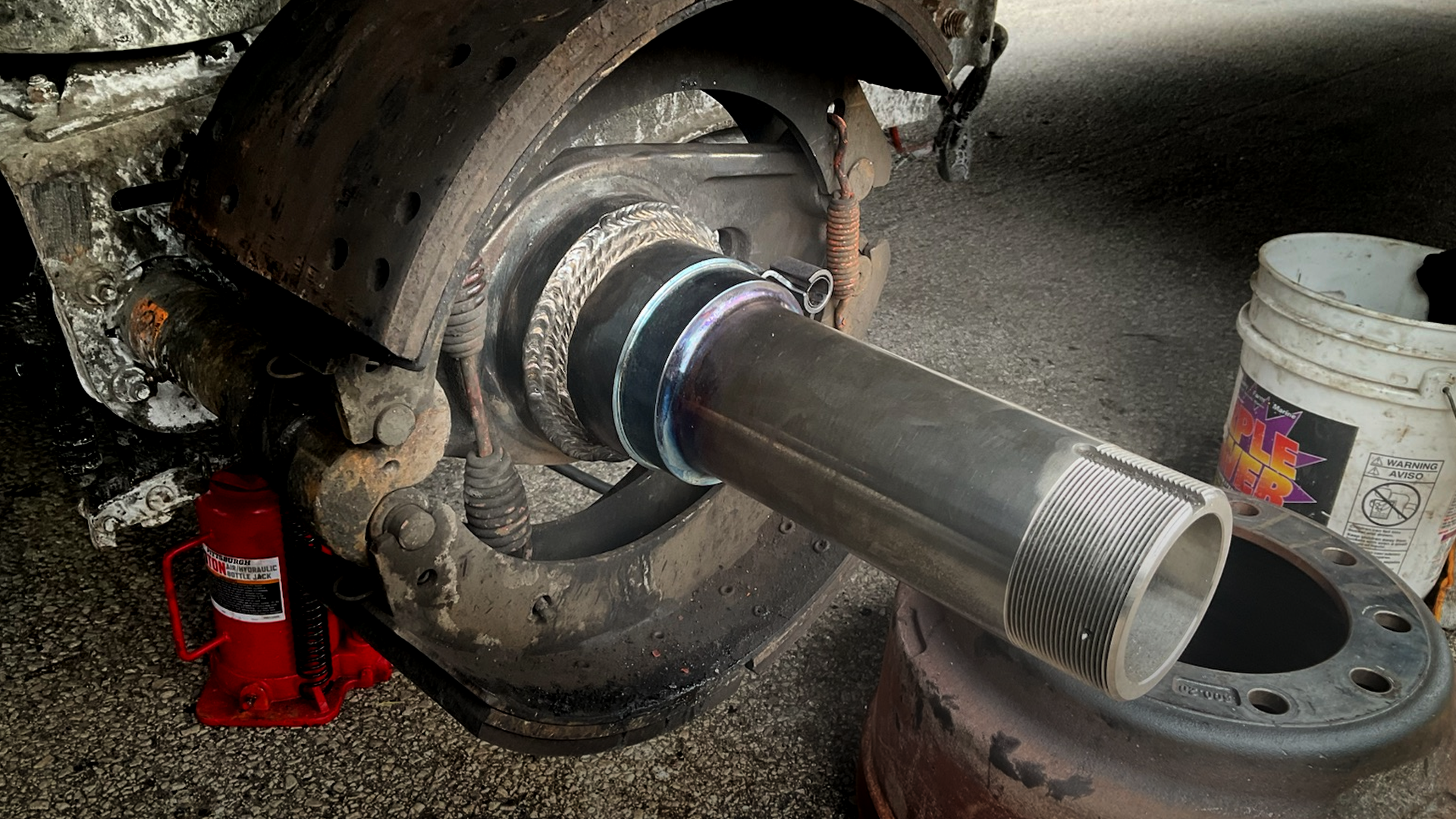 The wheel is removed from a semi-trailer axle, exposing the spindle and brake assembly while sitting on a gravel lot.