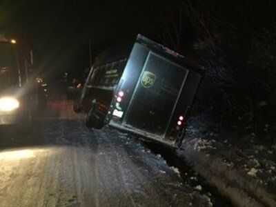 UPS truck tilted on its side on a snow-covered road at night, next to a truck with headlights on.
