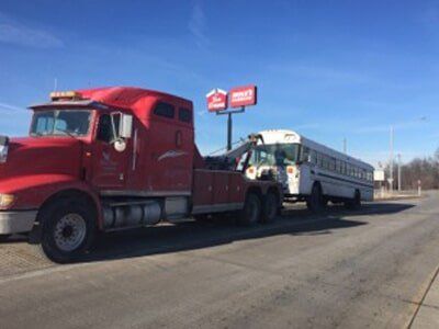A red tow truck towing a white school bus on a sunny road. A fast-food sign is in the background.