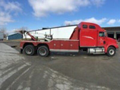 Red tow truck parked on an asphalt lot, with a blue-sky backdrop and a large, white building.