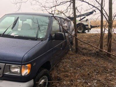Blue van stuck off the road, next to trees; a tow truck is in the background.