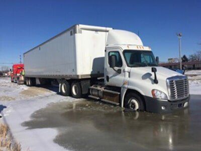 White semi-truck stuck in icy water next to a road on a clear, sunny day. A tow truck is visible in the background.