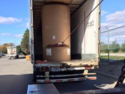 A large roll of brown paper inside an open truck trailer, secured by a strap. Outside, a paved lot and a fence are visible.