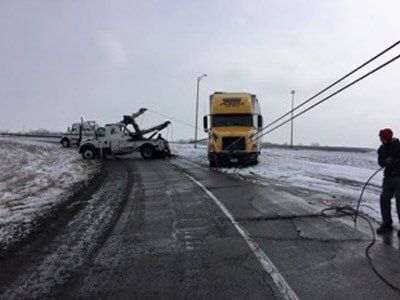 A tow truck attempts to pull a yellow semi-truck from snow-covered road. A man stands near the scene, holding a cable.