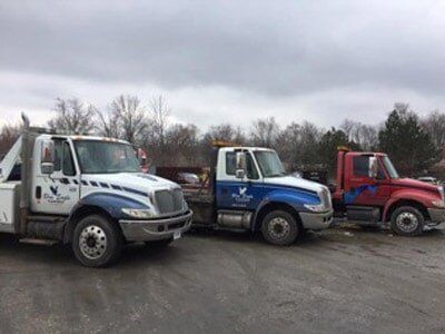 Three tow trucks parked on a gray asphalt lot under an overcast sky. The trucks are white, blue, and red.