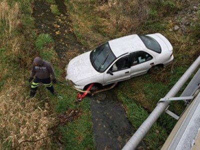A white car stuck in a ditch with a person in dark clothing pulling on a tow strap. 
