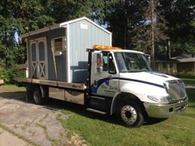 A tow truck carrying a small blue shed on its flatbed, parked on grass next to a driveway.