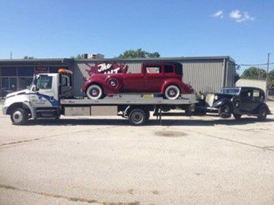 Tow truck carrying a red vintage car and towing a black vintage car on a sunny day.
