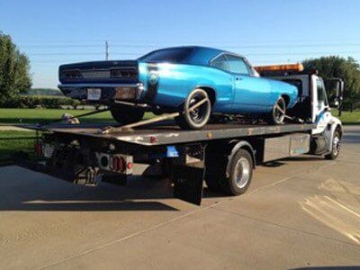 A light blue classic car is being towed on a flatbed truck on a paved surface, likely headed to a repair shop.