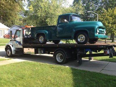 A teal vintage pickup truck is secured on the flatbed of a tow truck, parked on grass.