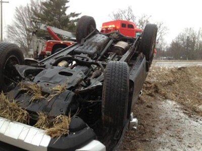 A pickup truck overturned on its side near a muddy roadside, with a tow truck in the background.