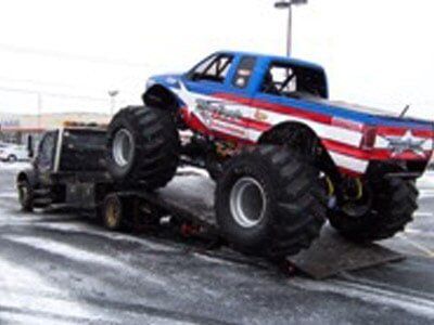 Monster truck with red, white, and blue design being loaded onto a black flatbed tow truck. Snow on the ground.