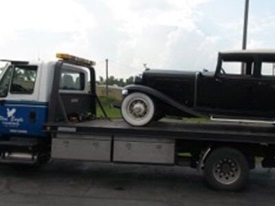 A black vintage car is being towed on a flatbed tow truck, likely for service. The tow truck is white and blue.