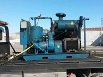 Blue industrial generator on a flatbed truck, secured with yellow straps, against a clear blue sky.