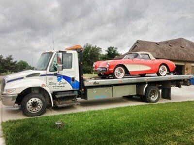 Red classic convertible car on a flatbed tow truck, possibly broken down, on a green lawn, under a cloudy sky.
