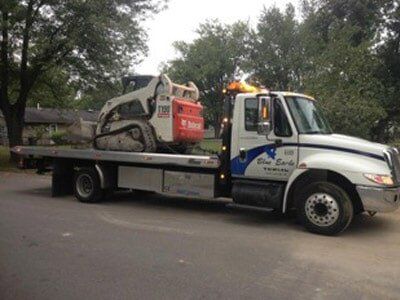 Tow truck transporting a white and orange Bobcat skid steer loader on a street. The truck is white with blue accents.