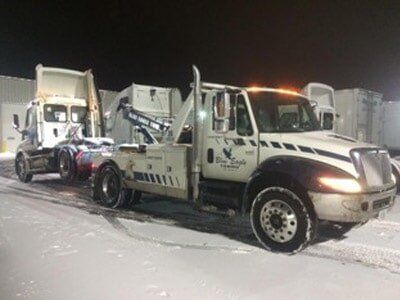 A tow truck hauling a semi-truck on a snowy night. The tow truck is white with blue and black accents.