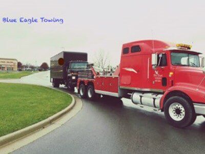 Red tow truck towing a dark truck on a wet road; Blue Eagle Towing logo in the background.