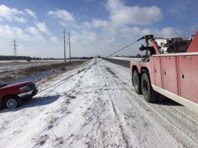 A red tow truck pulling a maroon truck out of snow-covered road. Power lines are visible in the background.