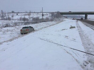 A car stuck in snow beside a highway on a snowy day. Another vehicle is on the road under an overpass.