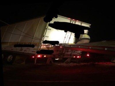 Semi-trailer truck tilted on its side at night with a worker visible; the trailer lights are on.