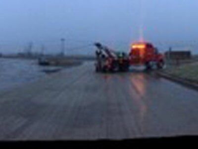 Tow truck with flashing lights towing a disabled vehicle on a wet road at dusk.