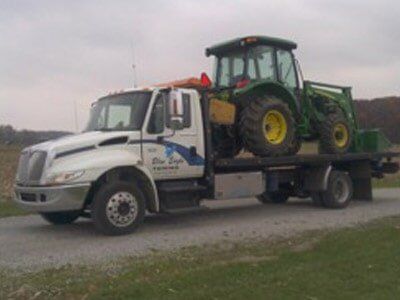White tow truck with a John Deere tractor on its flatbed, parked on a gravel road. The sky is overcast.