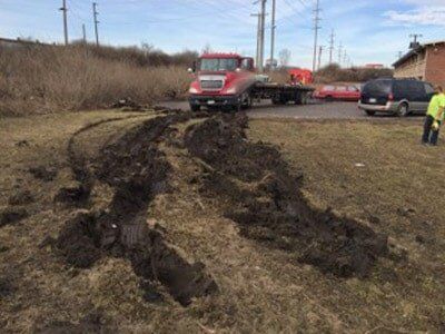 A red flatbed truck stuck in muddy ground. Deep tire tracks lead toward the truck. A person in a yellow vest stands nearby.