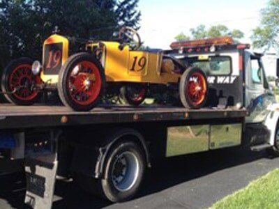 Yellow vintage race car with number 19 on a tow truck. The car has red wheels and is outdoors.