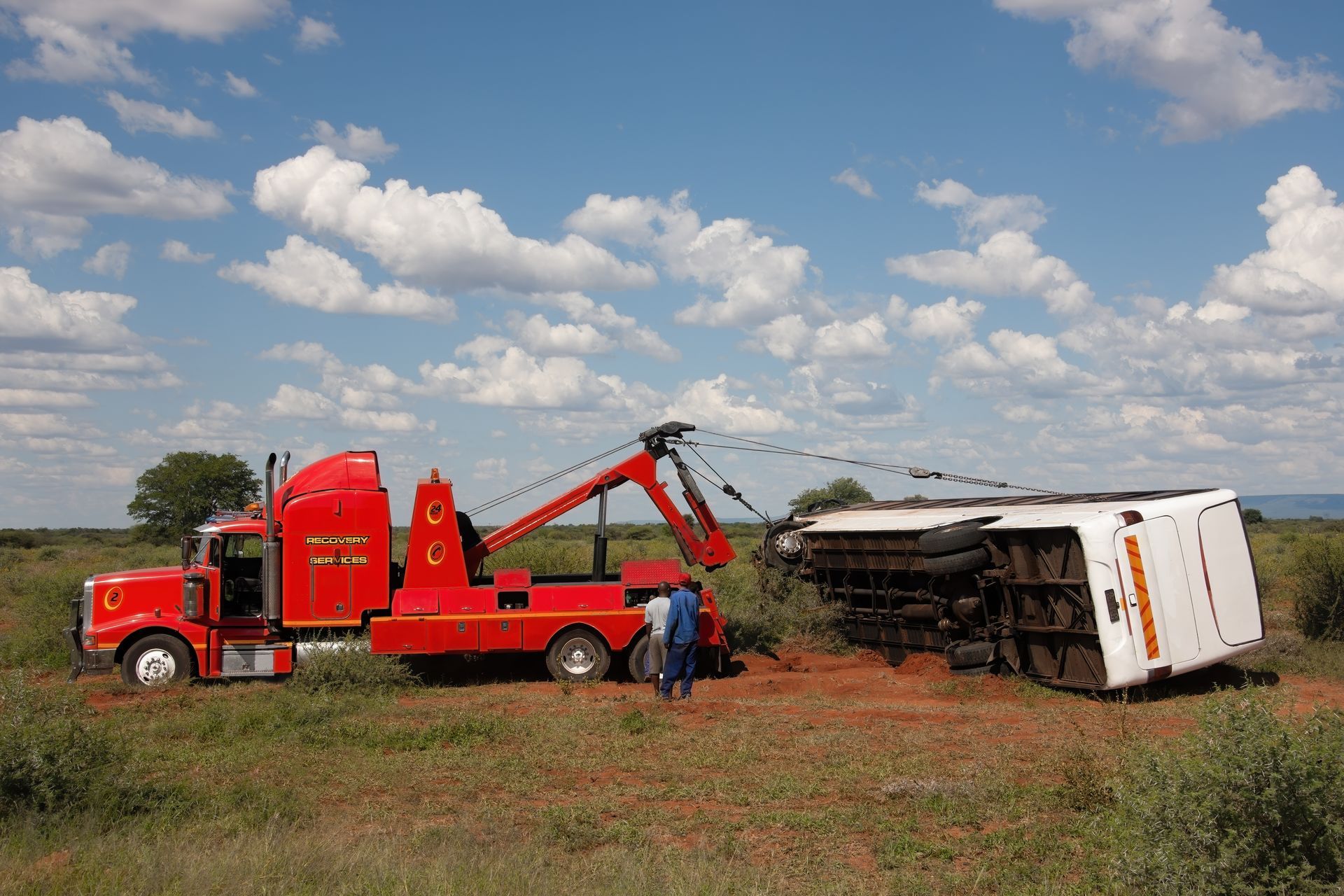 A heavy-duty towing truck with a crane righting a rolled-over coach bus in a rural area.