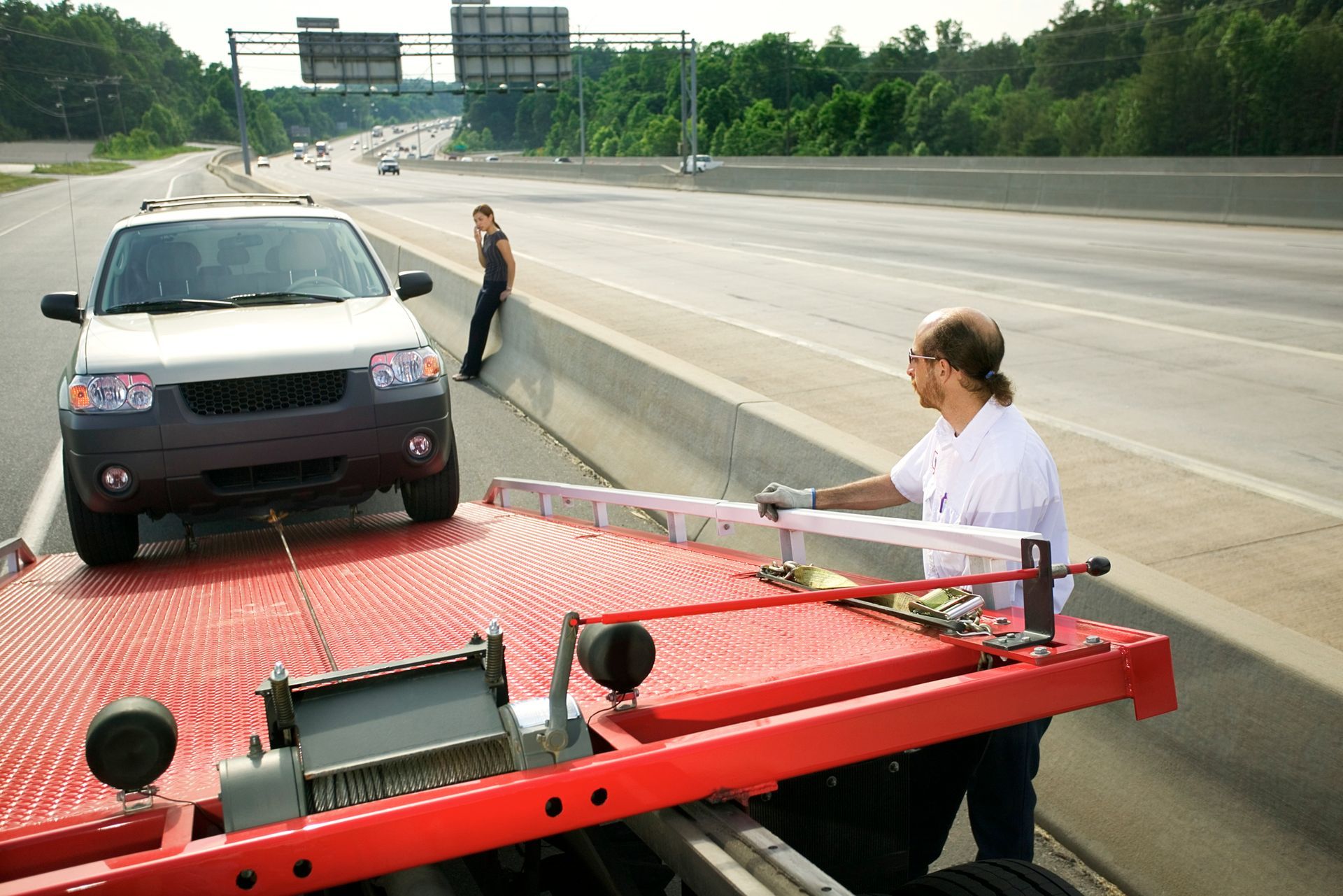 SUV being loaded onto a red tow truck on highway shoulder for roadside assistance service.