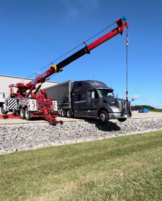 A large red tow truck lifting a black semi-truck's front end on a sunny day. The scene is outdoors near a building.