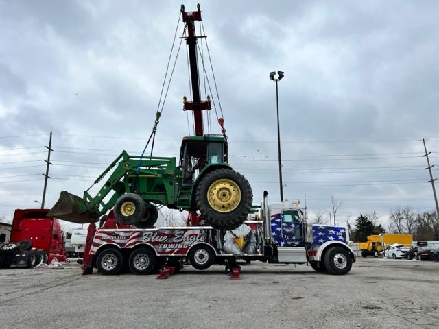 A large tow truck lifting a green John Deere tractor with a front loader, set in a parking lot under a cloudy sky.