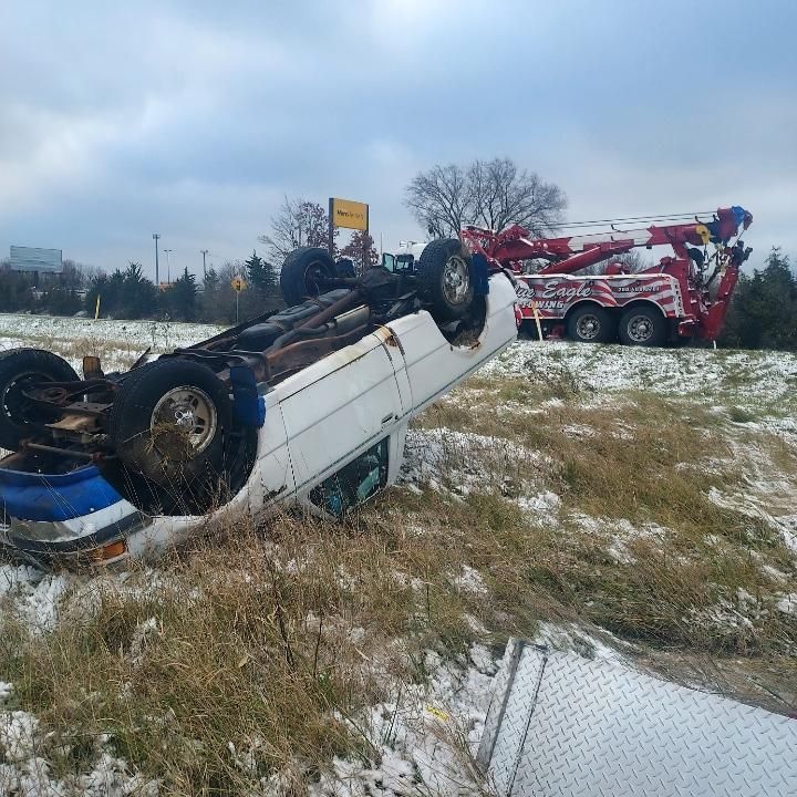 A white van overturned on its side in a snowy ditch, next to a red tow truck on a cloudy day.