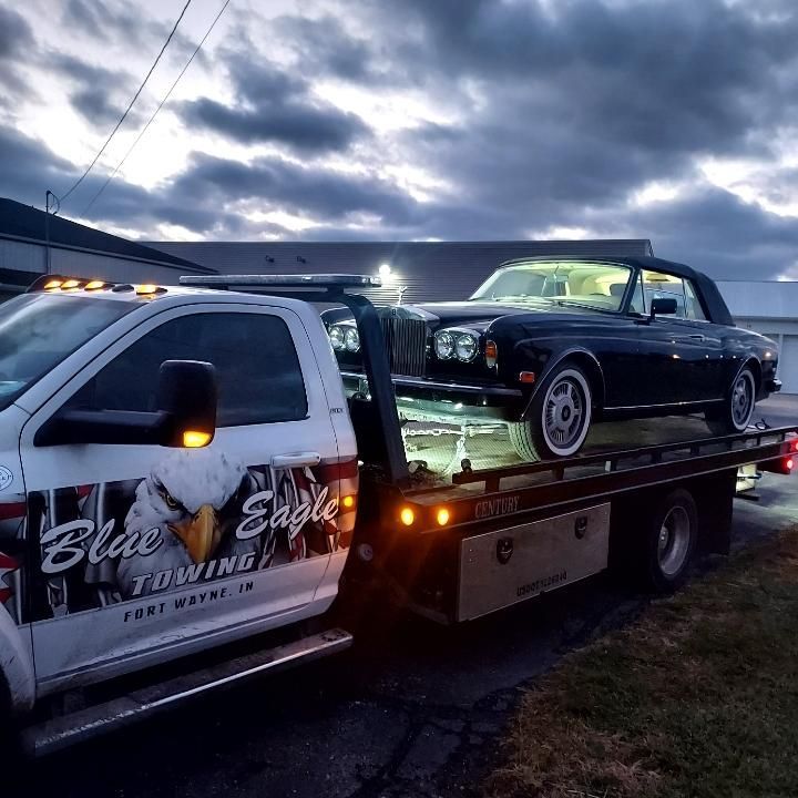 A black vintage convertible being towed on a flatbed truck by Blue Eagle Towing in Fort Wayne, IN, at dusk.