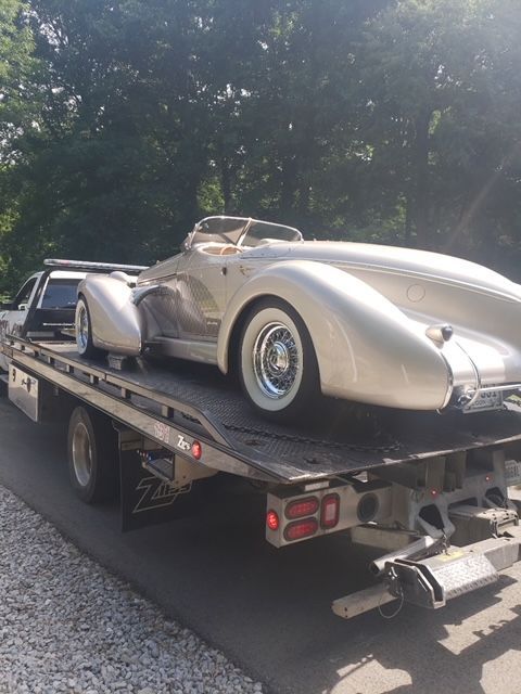 A silver classic car with long fenders is being transported on a flatbed tow truck on a paved road.