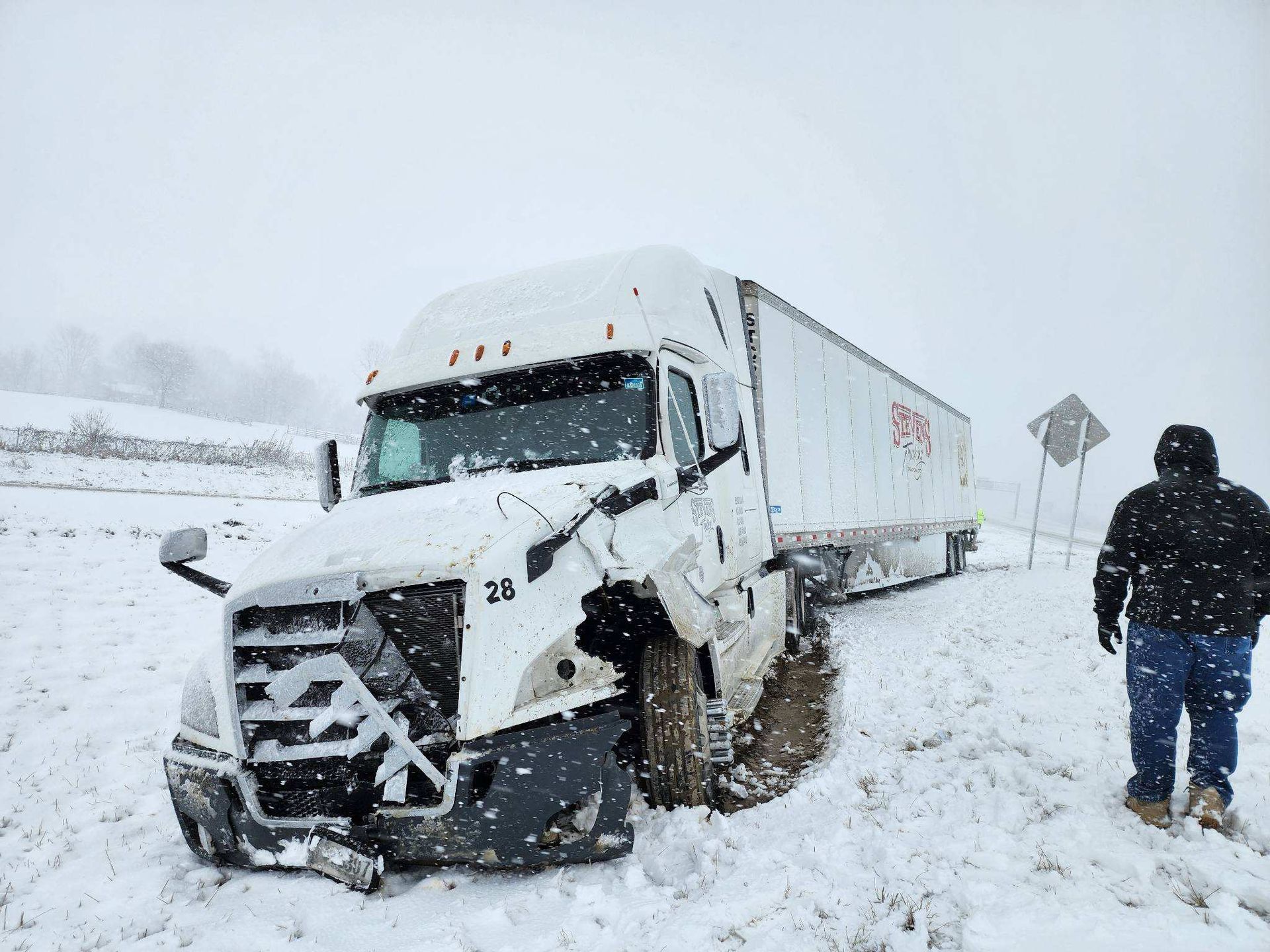 Semi-truck stuck in snow on side of road; a person walks away. Snowy, overcast day.