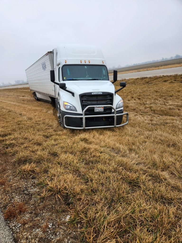 A white semi-truck is stuck off the road in a field of dry grass. Overcast sky, brown and beige tones.
