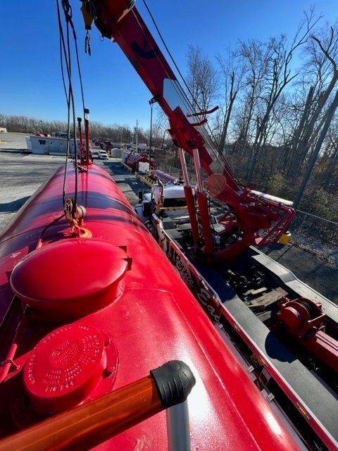 Red tanker truck being loaded or unloaded by a red crane; outdoors on a sunny day.